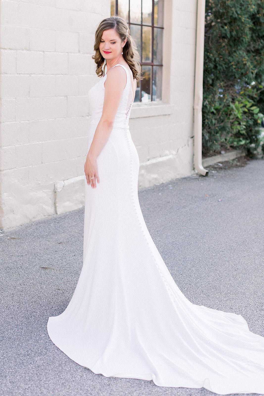 Bride in a white wedding dress with a long train, standing outside near a brick wall and window.