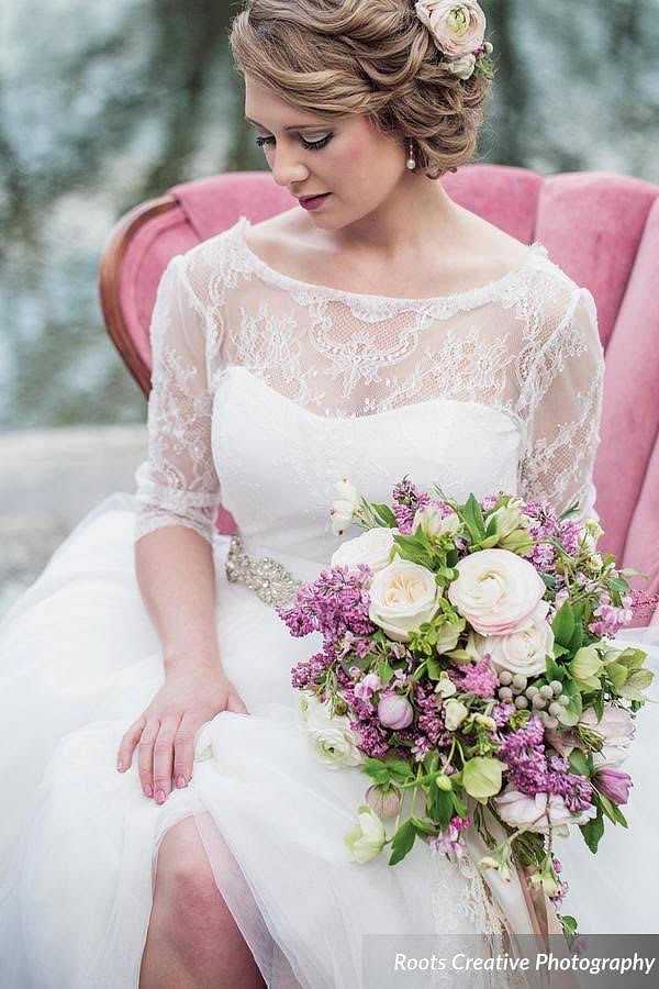 Bride in lace dress holding floral bouquet, seated on pink chair, with serene outdoor setting.