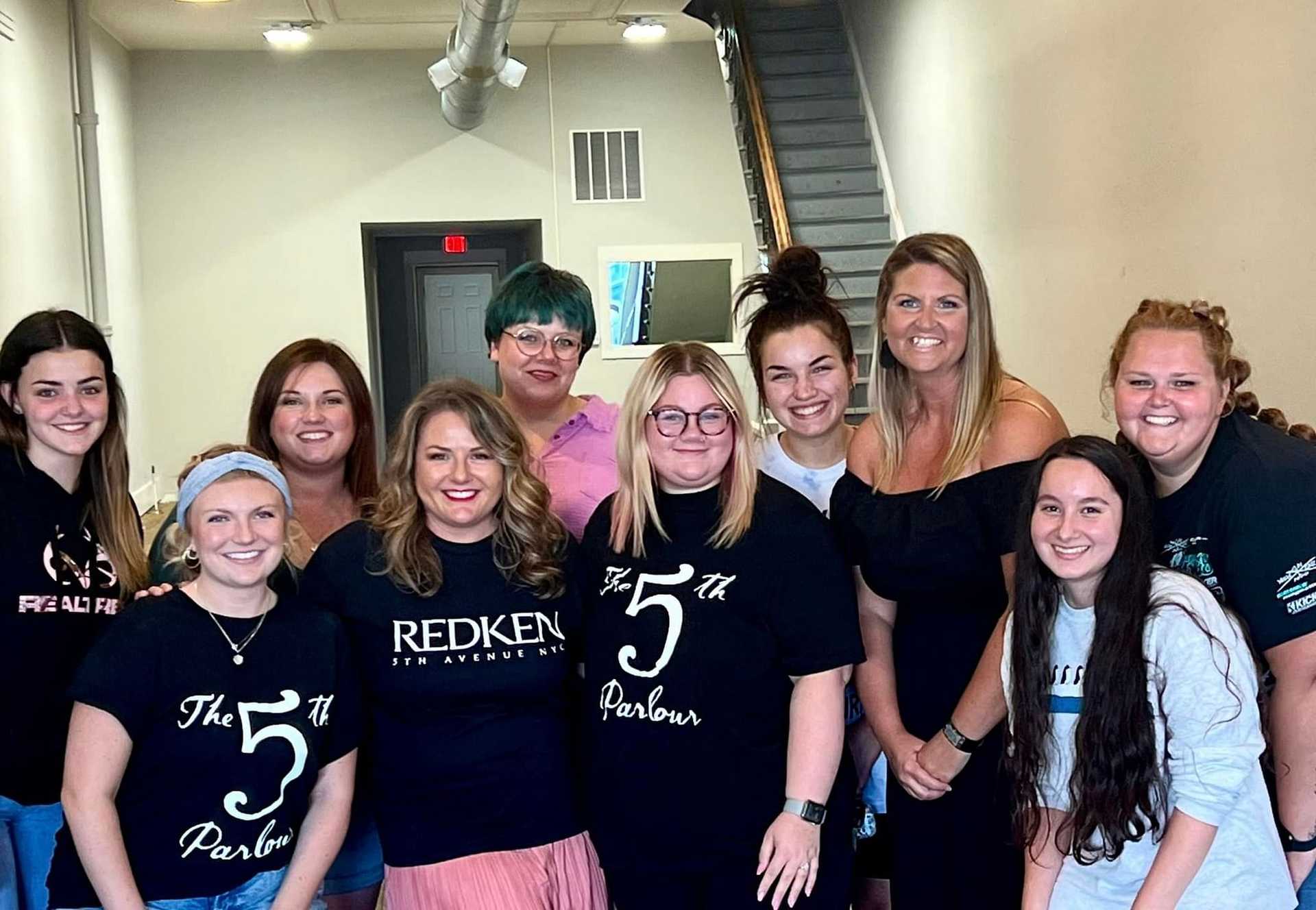 Group of ten women smiling inside a hair salon, some wearing shirts with "The 5th Parlour" logo.