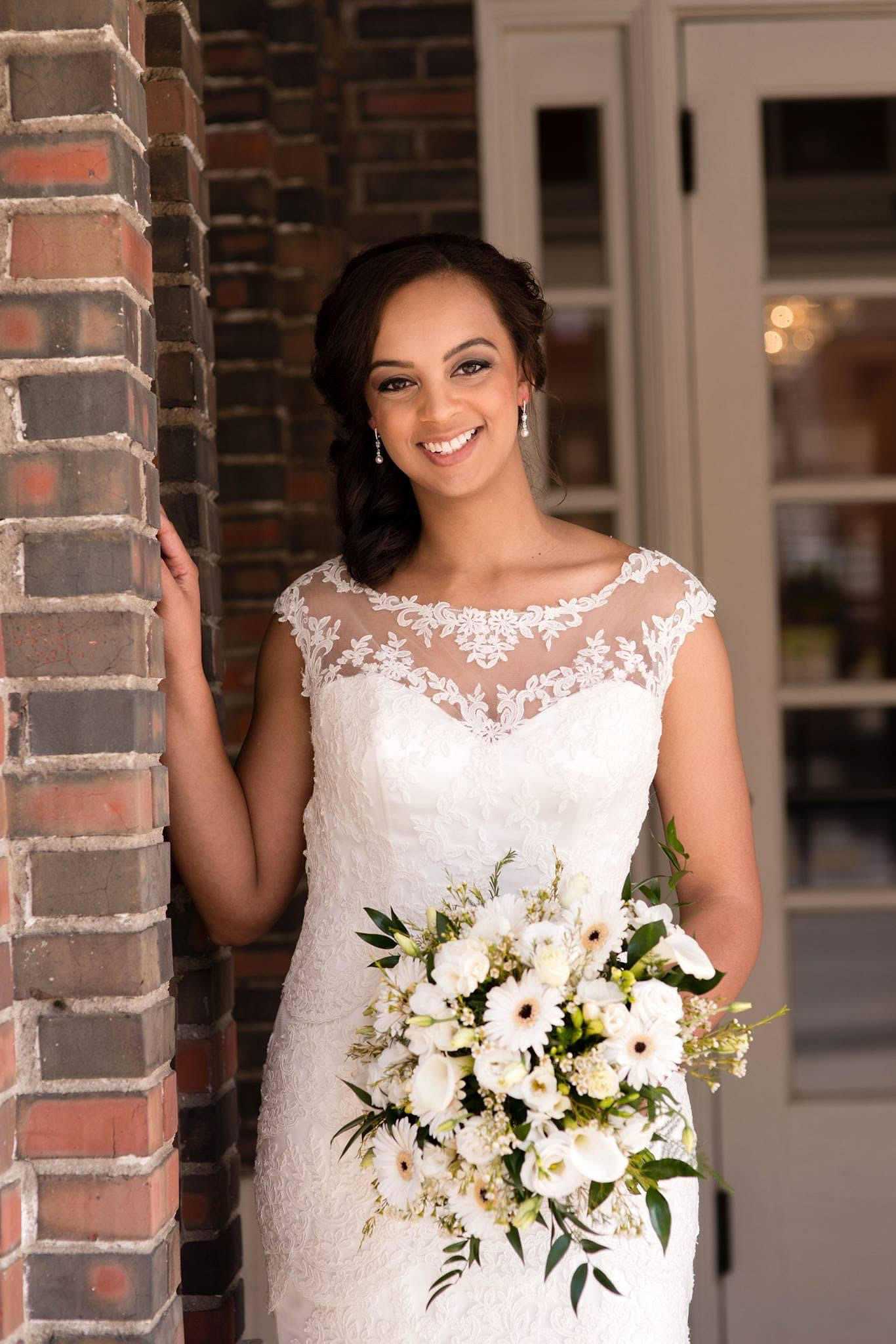 Bride in lace wedding gown holding white and green bouquet, smiling against brick doorway backdrop.